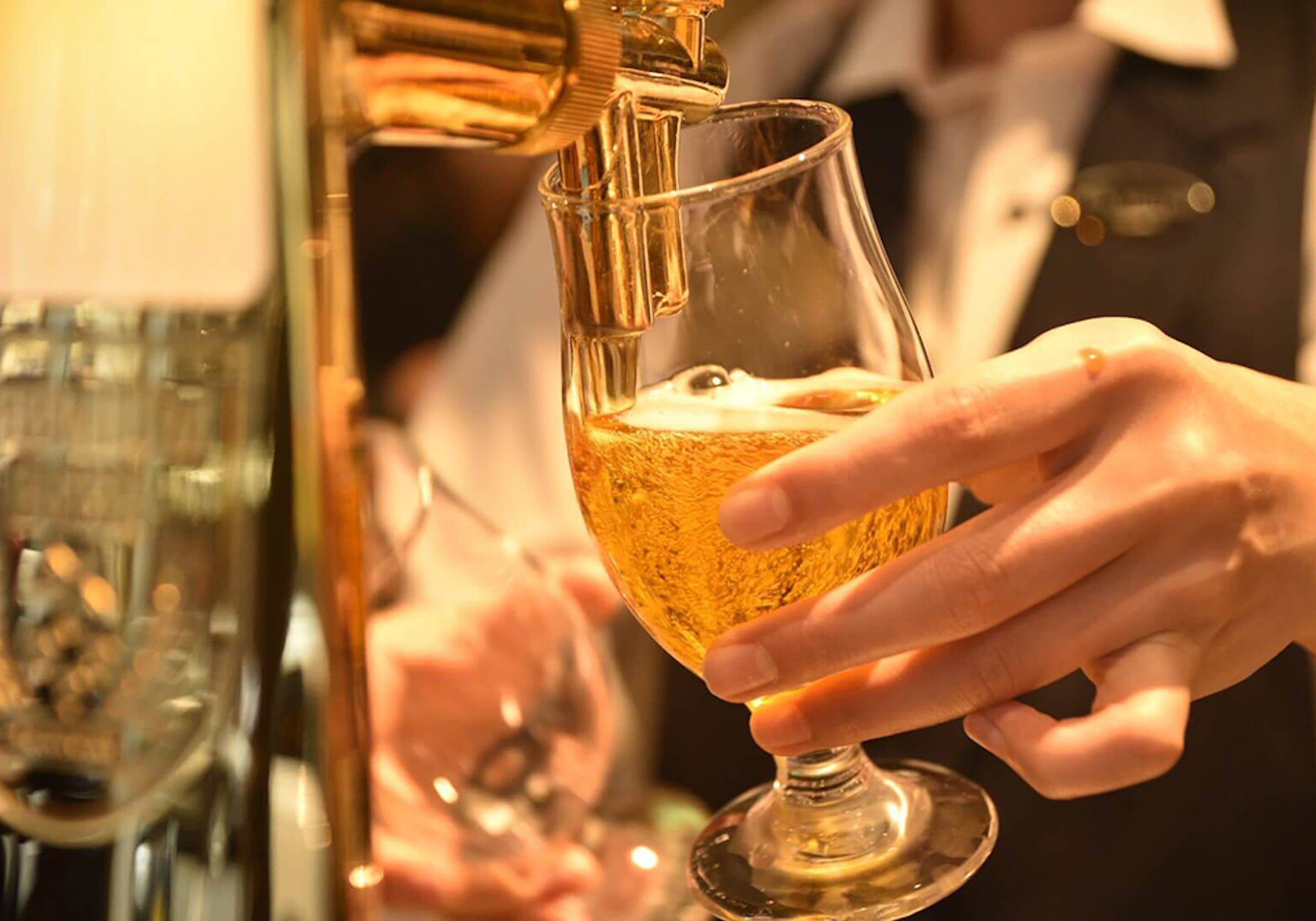 Una mano sosteniendo un vaso con un líquido dorado, probablemente un cóctel, que se está sirviendo desde un grifo.
