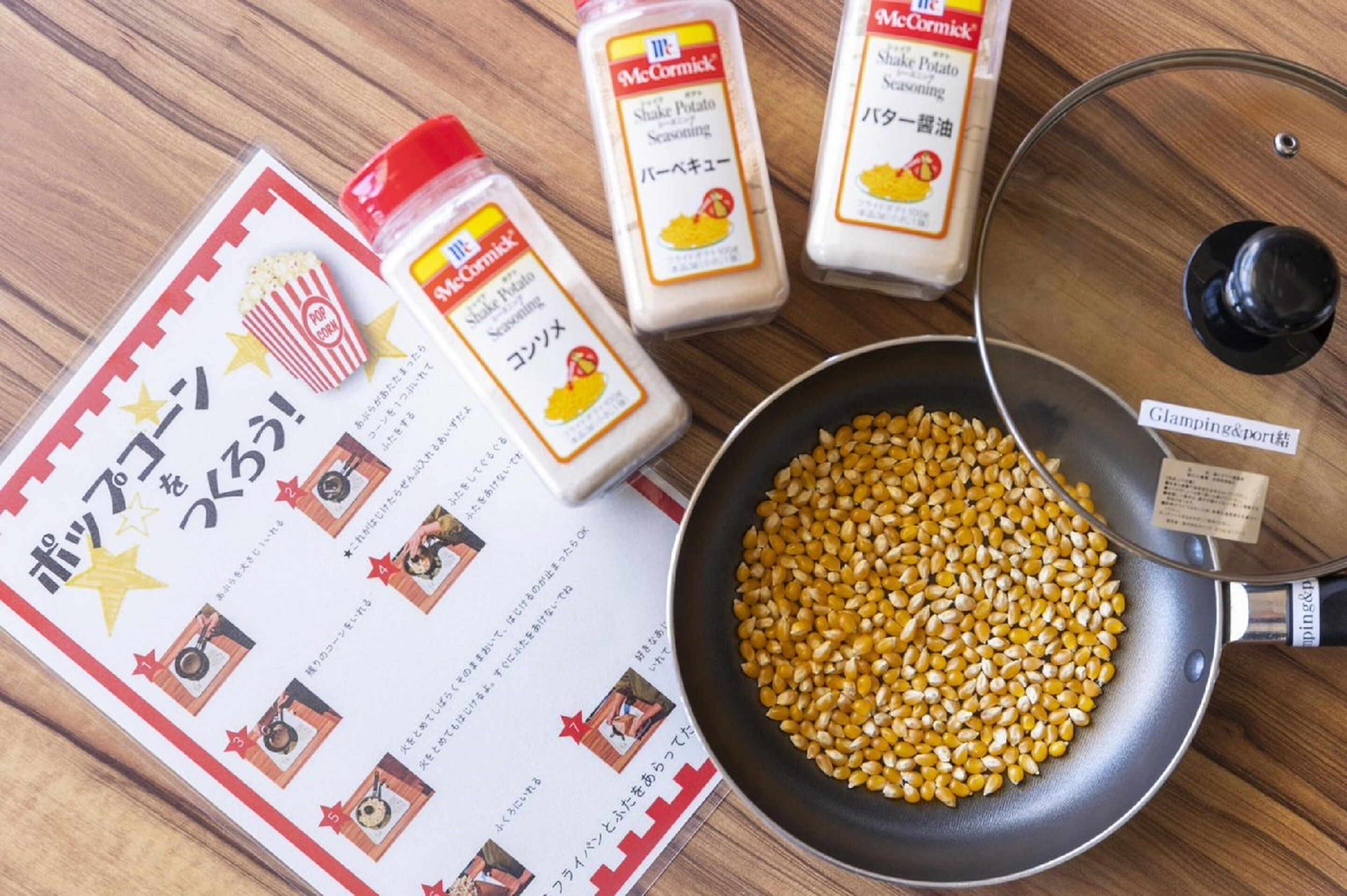A cooking pan with corn kernels, a popcorn container, and seasoning bottles on a wooden surface.
