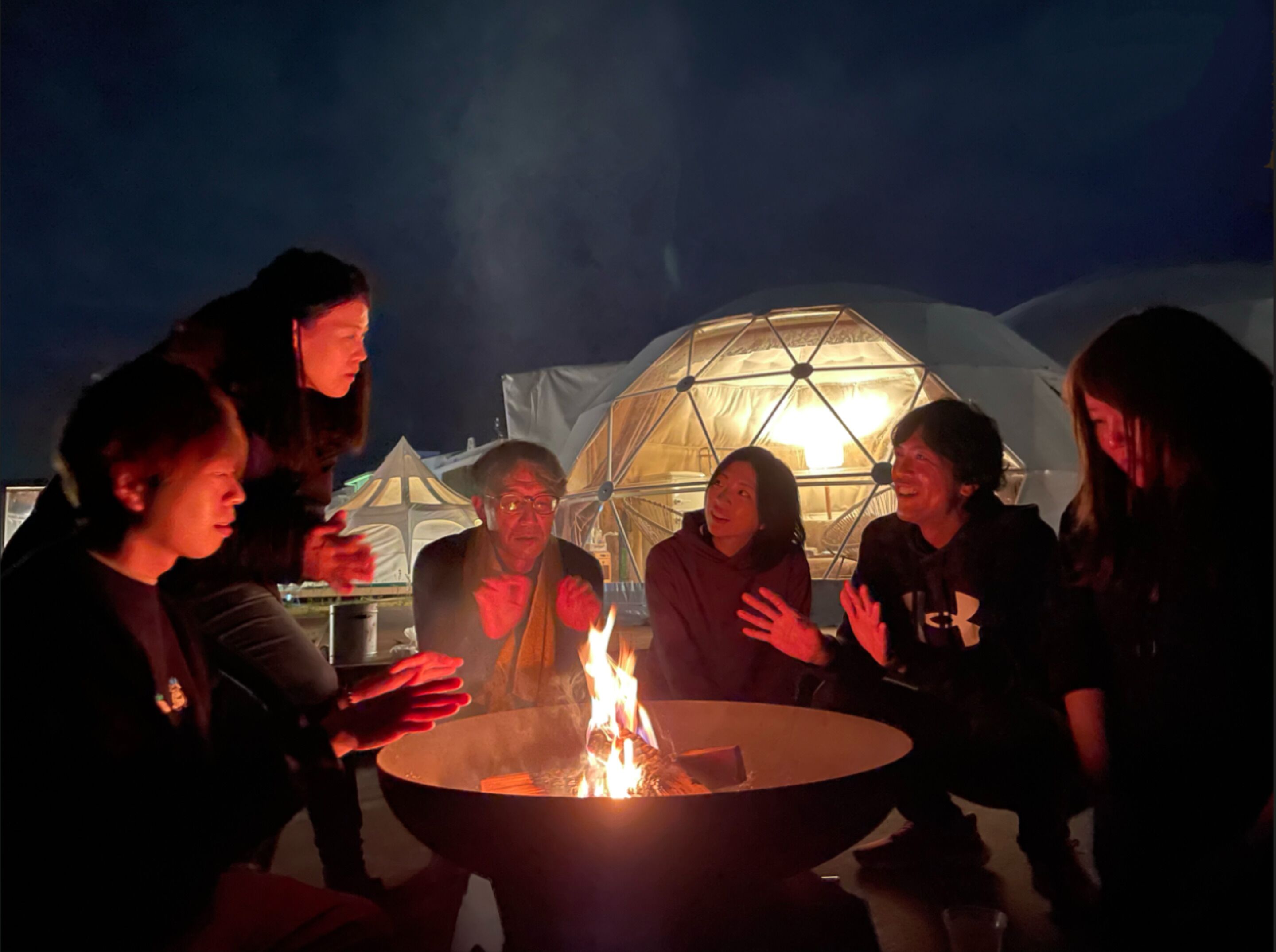 Eine Gruppe von Menschen versammelt sich um eine Feuerstelle, im Hintergrund eine geodätische Kuppel.