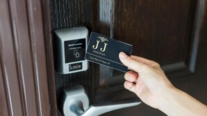 Eine Hand hält eine schwarze Karte mit dem Schriftzug "JJ RESIDENCE" und "Gemütlicher Ort & Glückliches Leben". Im Hintergrund eine Tür mit digitalem Schloss und einem Schild mit der Aufschrift "Willkommens-Induktionszone".