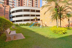 A playground with slides and swings, surrounded by a grassy area and a building with a curved facade.