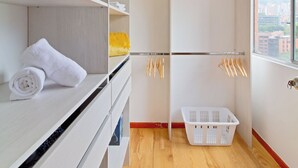 Une salle de bain moderne avec un plancher en bois, des étagères blanches où sont posées des serviettes, un panier à linge blanc et une vue sur les immeubles par une fenêtre.