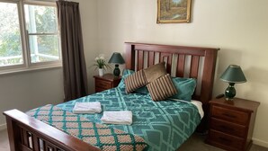 A neatly made bed with a patterned bedspread, two pillows, and two folded towels. There is a nightstand with a lamp and a small table with a plant. A painting hangs on the wall above the bed. - Family farm retreat, big outdoor alfresco areas, with emus (Barwidgee)