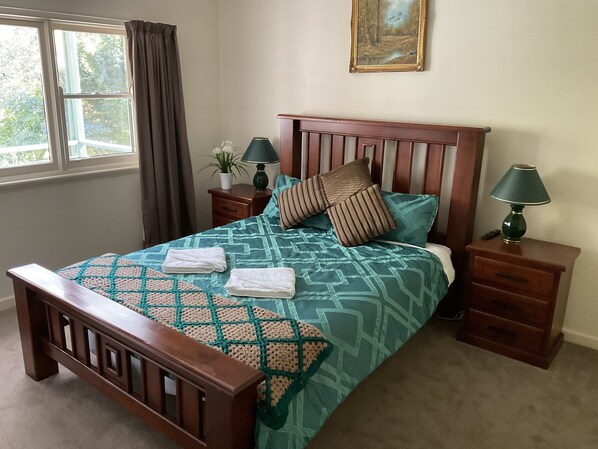 A neatly made bed with a patterned bedspread, two pillows, and two folded towels. There is a nightstand with a lamp and a small table with a plant. A painting hangs on the wall above the bed. - Family farm retreat, big outdoor alfresco areas, with emus (Barwidgee)