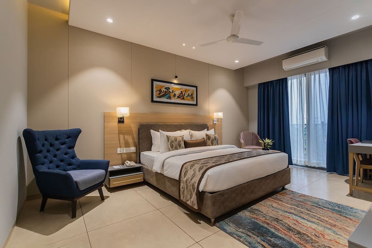 A modern hotel room with a large bed, a blue armchair, a desk, and a ceiling fan.