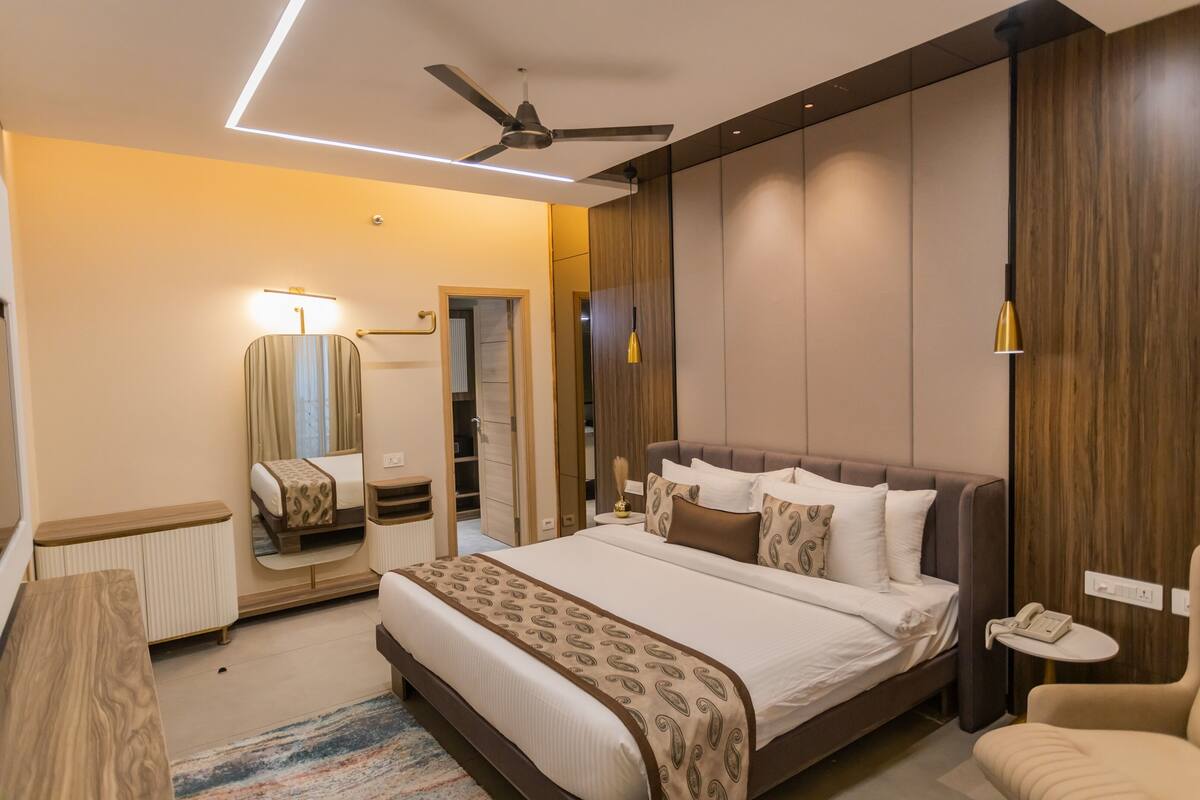 A modern hotel room with a large bed, a ceiling fan, a bedside table, a nightstand, a lamp, a mirror, a wardrobe, and a small round table with a phone.