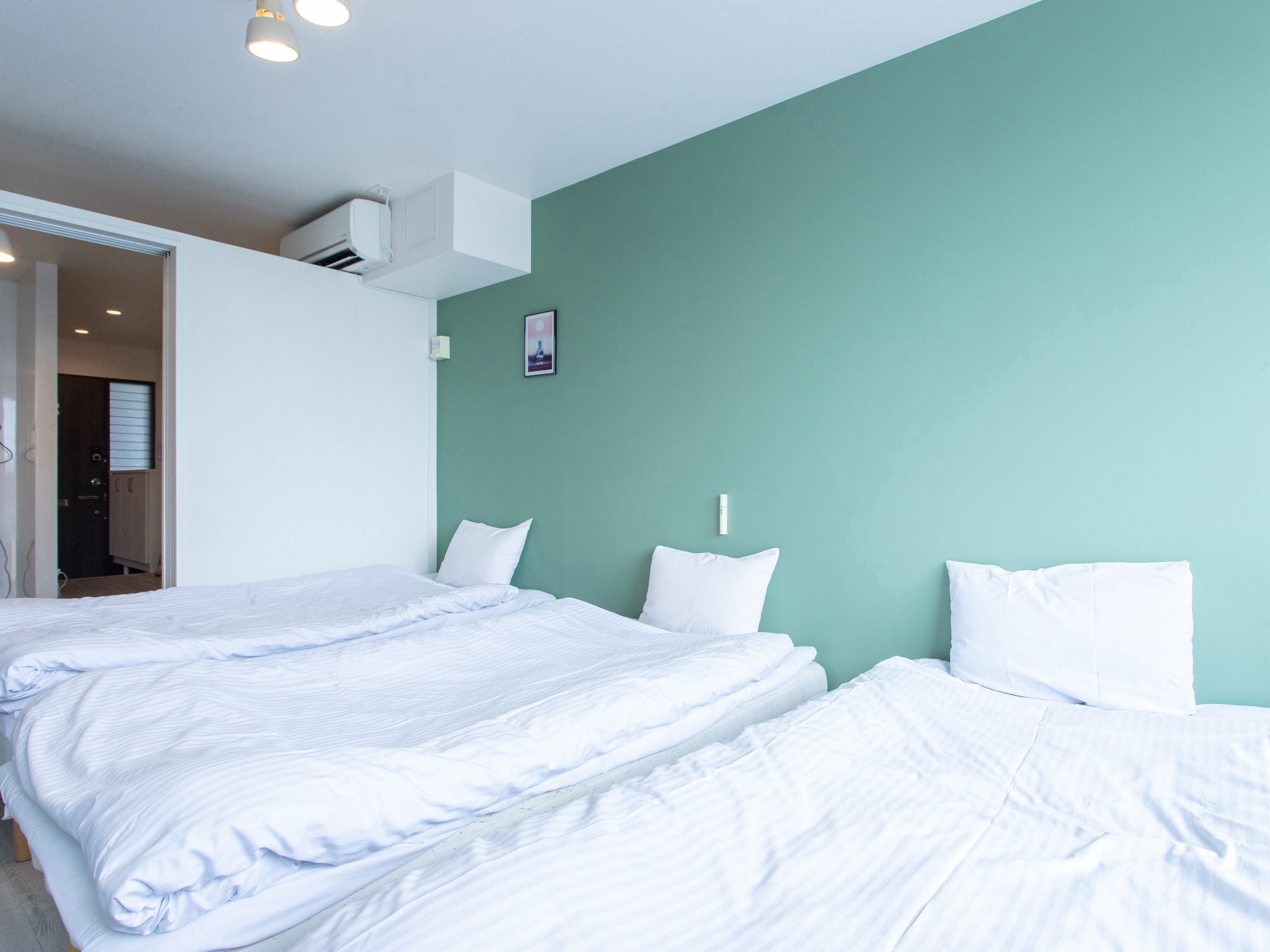 Hotellihuone, jossa on kaksi sänkyä, valkoiset vuodevaatteet, turkoosinsävyinen korosteseinä ja ovi, joka johtaa kylpyhuoneeseen.