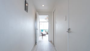 Um corredor com piso de madeira e paredes brancas, que leva a um quarto com cama e escrivaninha.