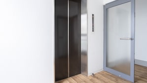 Elevador moderno com acabamento metálico e porta de vidro, próximo a uma porta com puxador metálico.