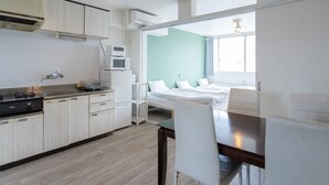 Cozinha compacta com armários brancos, fogão e micro-ondas. No quarto adjacente, há uma mesa de jantar com cadeiras e uma cama com roupa de cama branca.