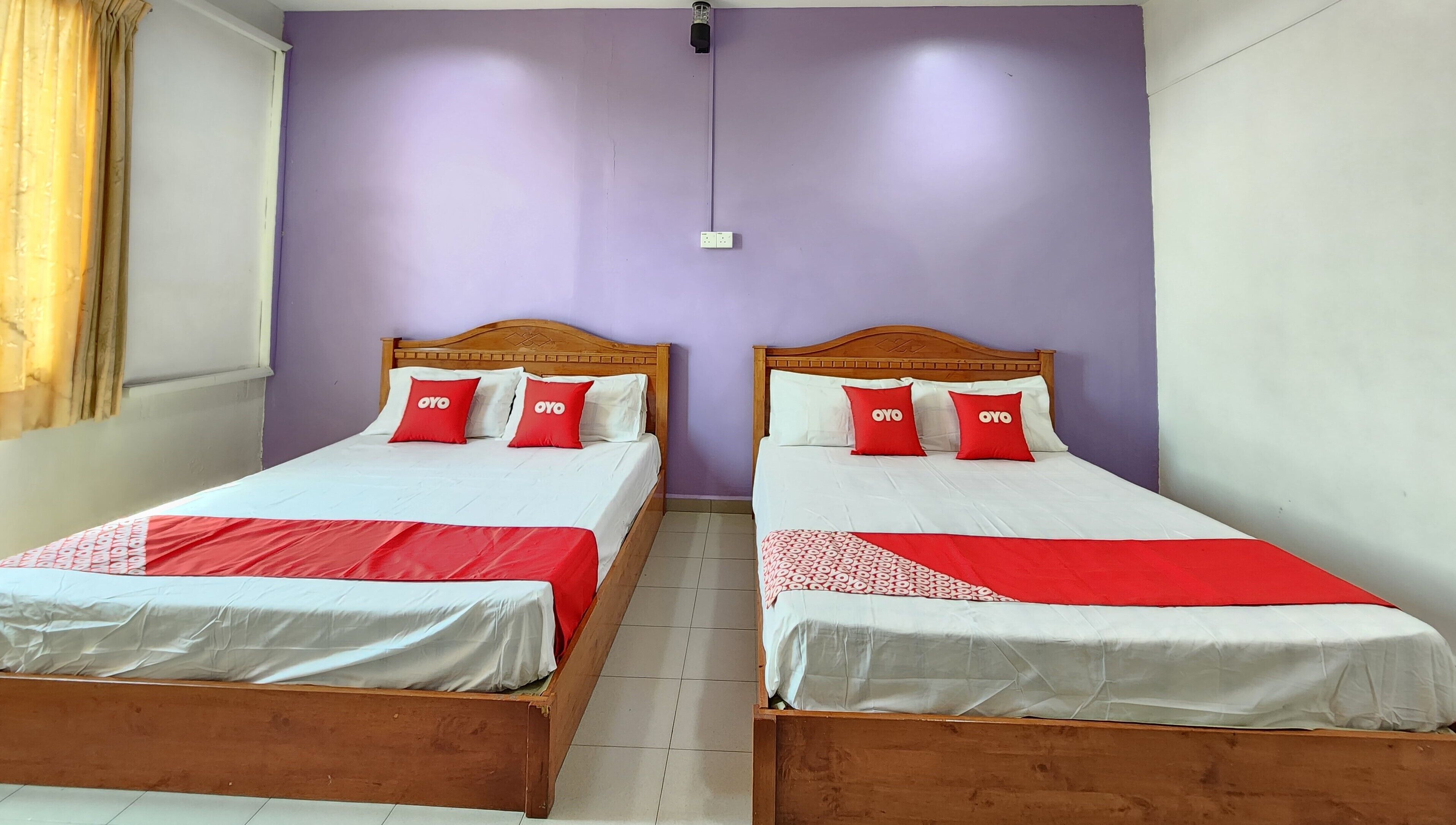 Two wooden beds with white and red bedding, a purple wall, and a window with curtains.