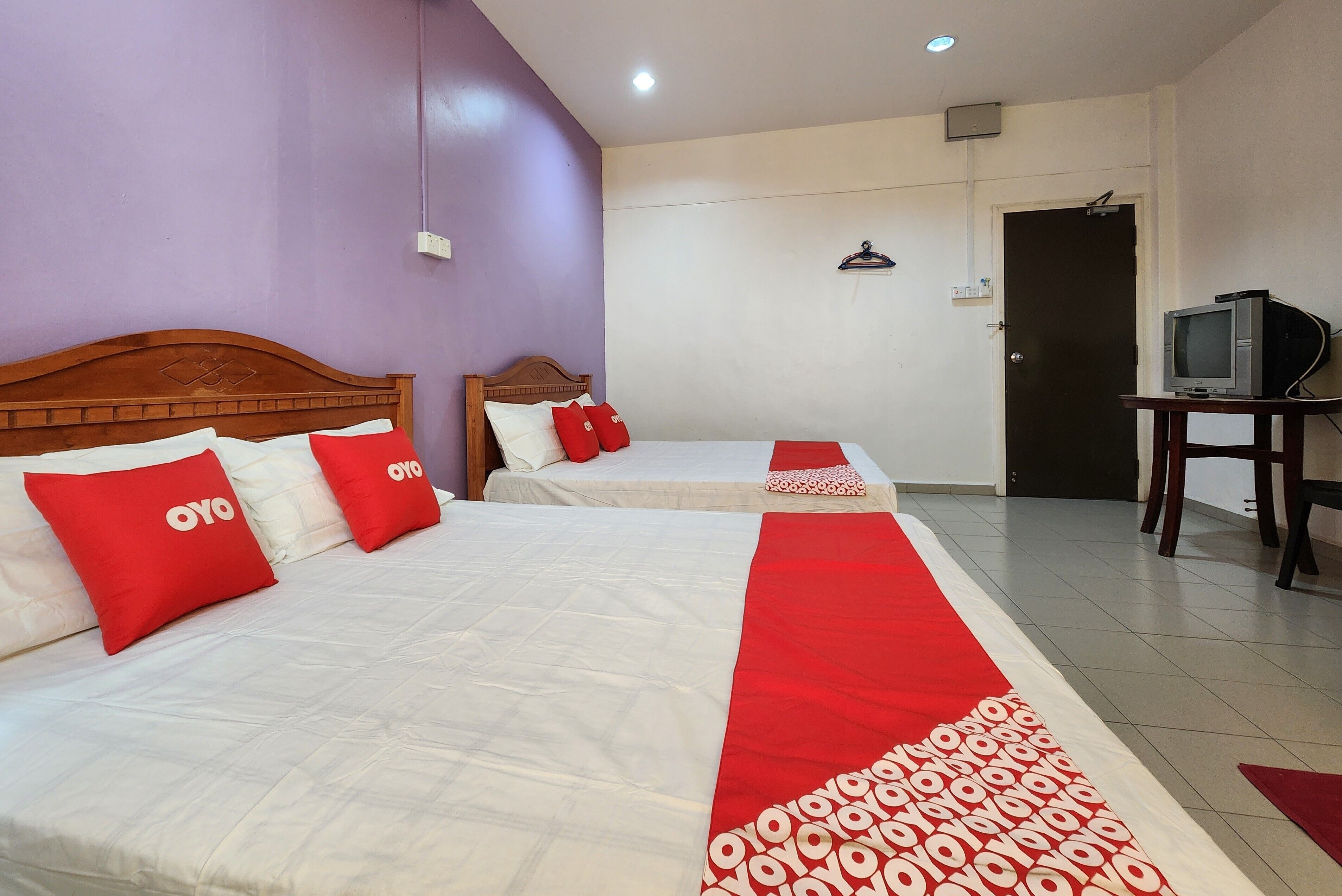 Una habitación de hotel con dos camas, cada una con ropa de cama roja y blanca, un corredor con estampado rojo y blanco y un televisor sobre una mesita.
