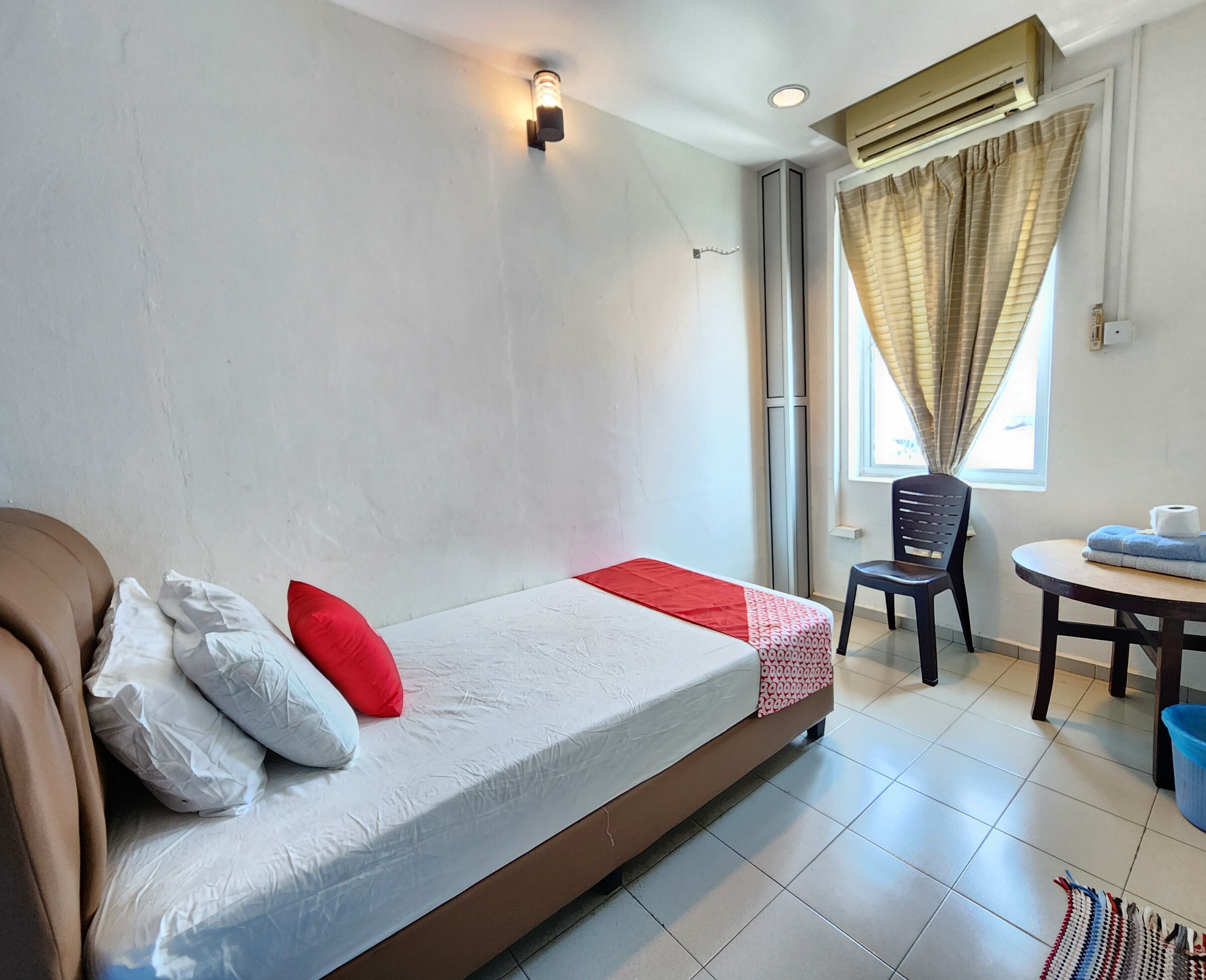 A single bed with a white mattress, red and white pillows, a red blanket, and a wall-mounted light fixture. There is a window with curtains, a chair, and a small table with a towel and a trash bin.