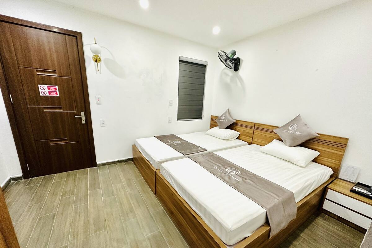 A bedroom with a wooden bed, two pillows, a nightstand, a wall-mounted fan, a window with blinds, and a door with a sign.