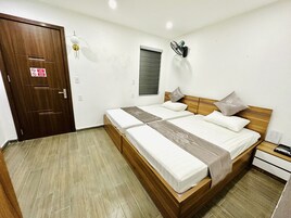 Un dormitorio con cama de madera, dos almohadas, una mesita de noche, un ventilador de pared, una ventana con persianas y una puerta con un cartel.