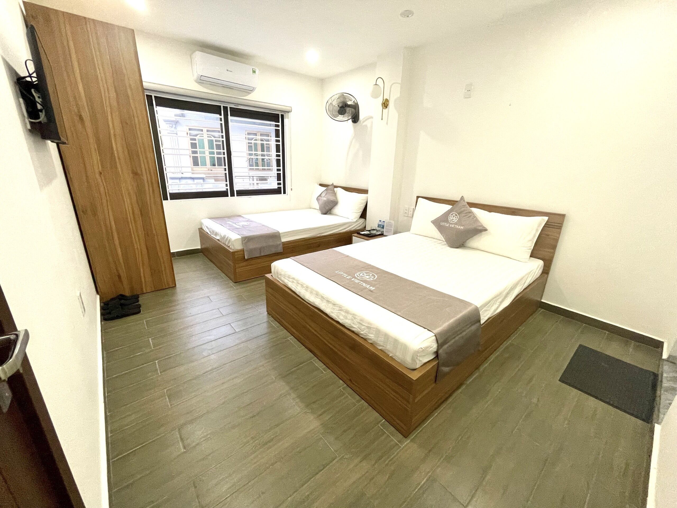 A hotel room with two beds, a window with blinds, an air conditioning unit, a wall-mounted fan, and a door.