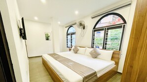 A hotel room with a large bed, a standing fan, and a window with a view of greenery. - Little Vietnam Hotel - Cat Ba (Hai Phong)