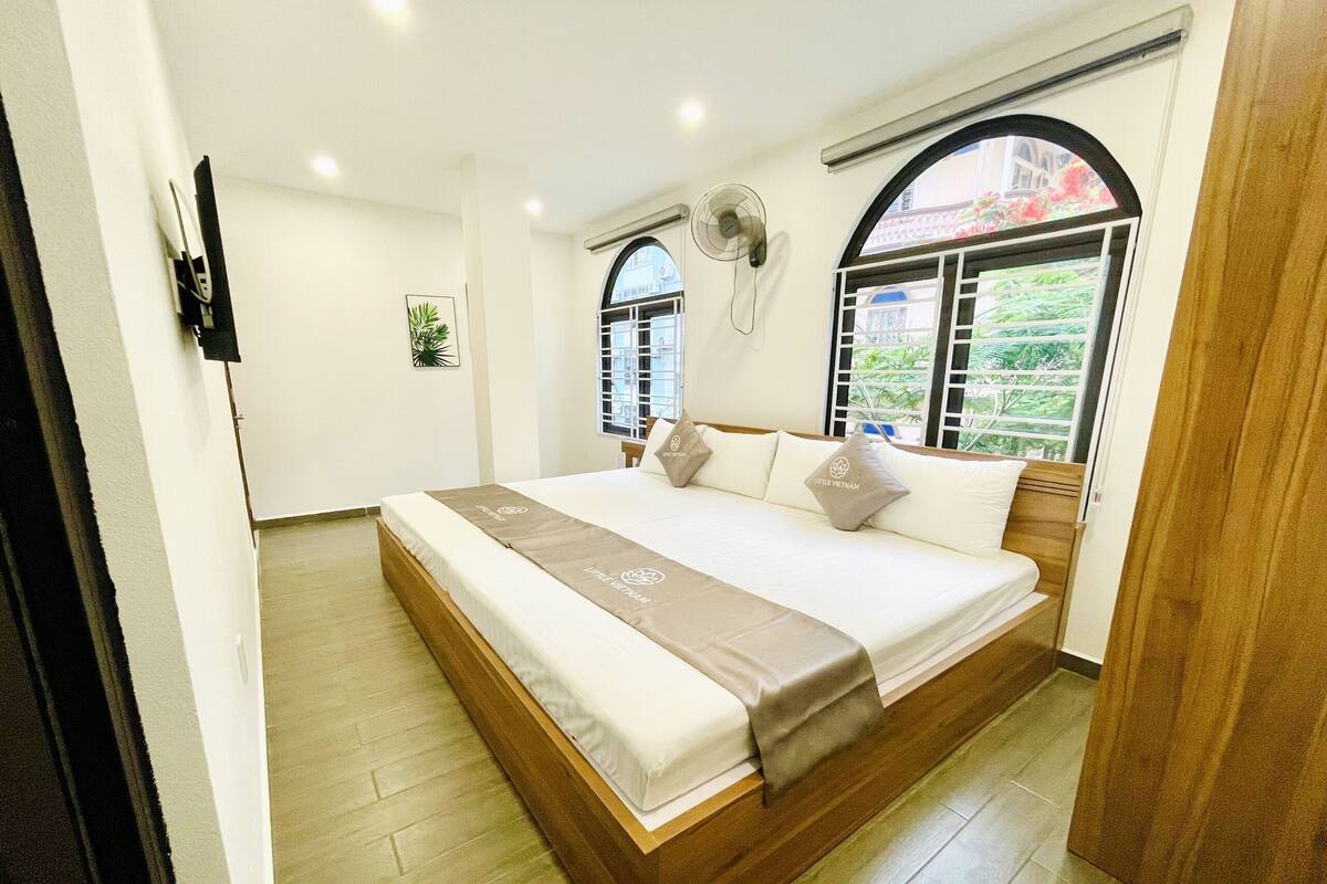 A hotel room with a large bed, a standing fan, and a window with a view of greenery.