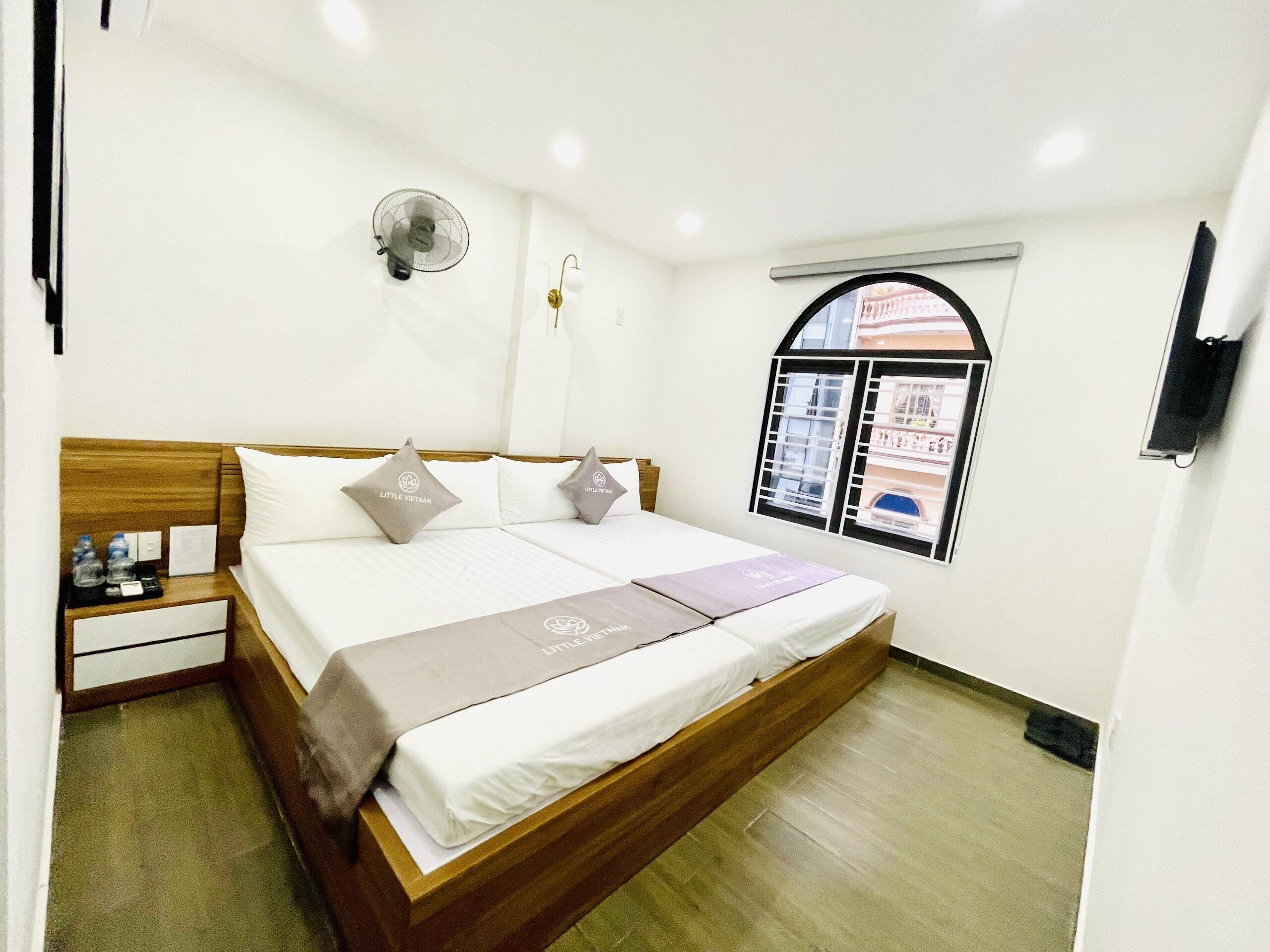 A hotel room with a double bed, a bedside table with water bottles, a wall-mounted fan, and a window with a view of a building.