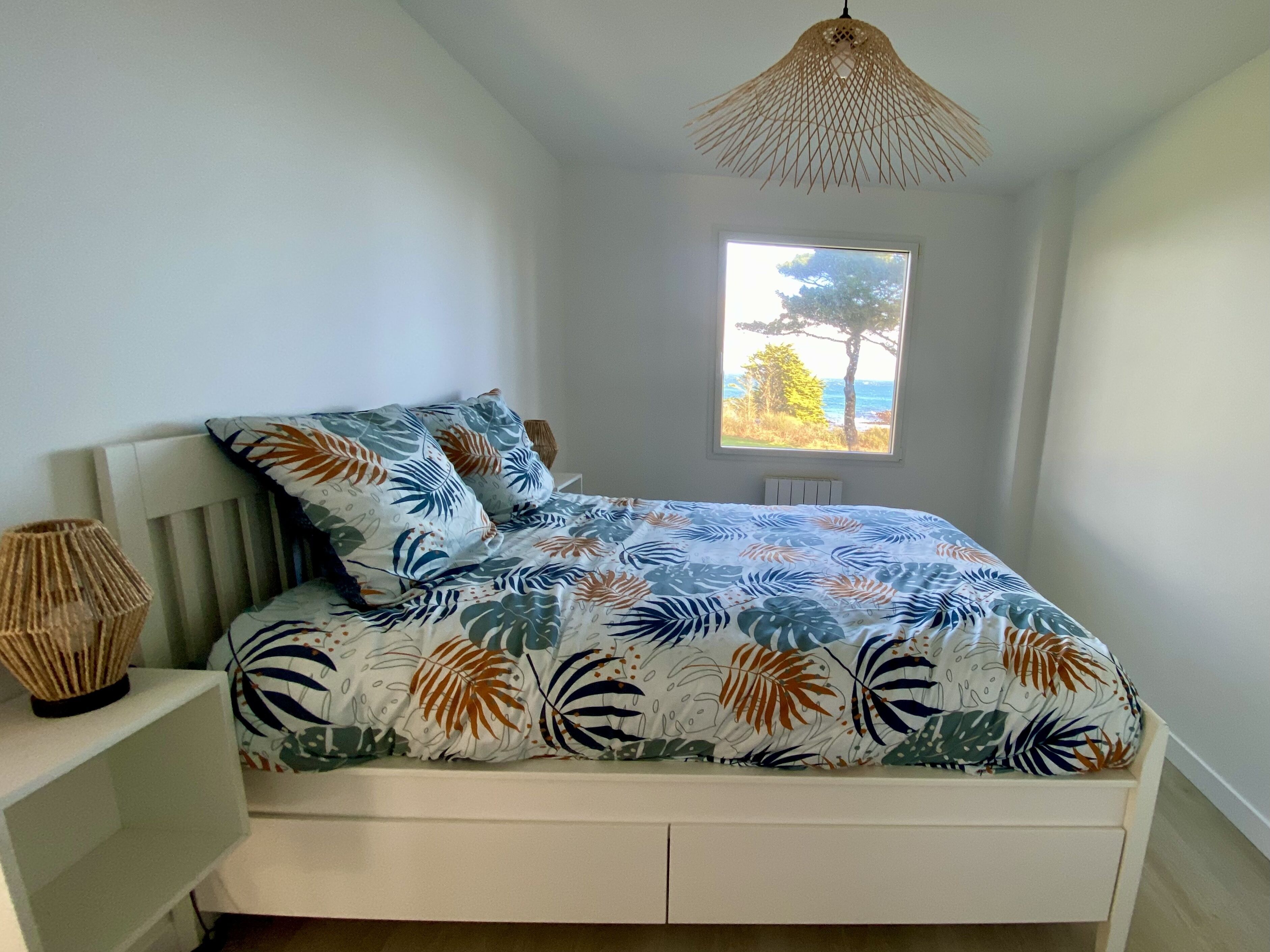 A bedroom with a bed, a window with a view of trees and the sea, a woven lamp, and a white bedside table.