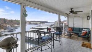 A balcony with a view of a river and boats, a ceiling fan, a grill, and a sofa with cushions. - Lighthouse Retreat – Pool, Swim Dock and More! (Lake Ozark)