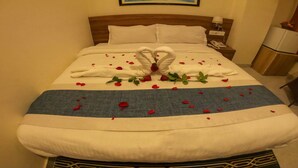Una cama bien hecha adornada con pétalos de rosa y una escultura de toalla, una mesita de noche con una lámpara y un refrigerador en la esquina.