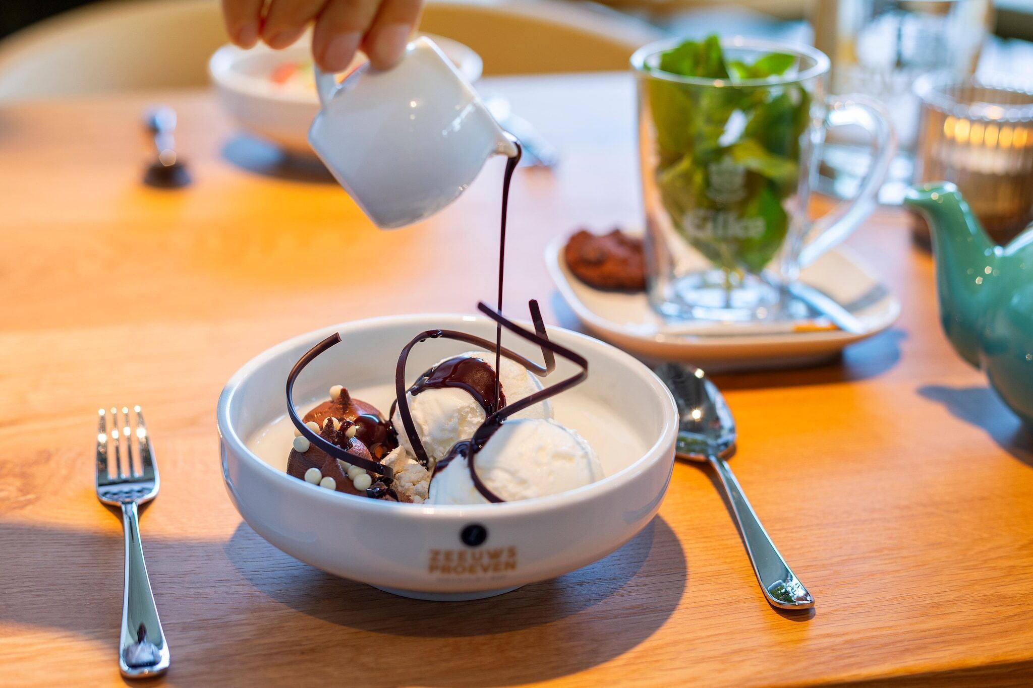 Een kom ijs met chocoladesaus en noten, een glas ijsthee, en een theepot op een houten tafel.
