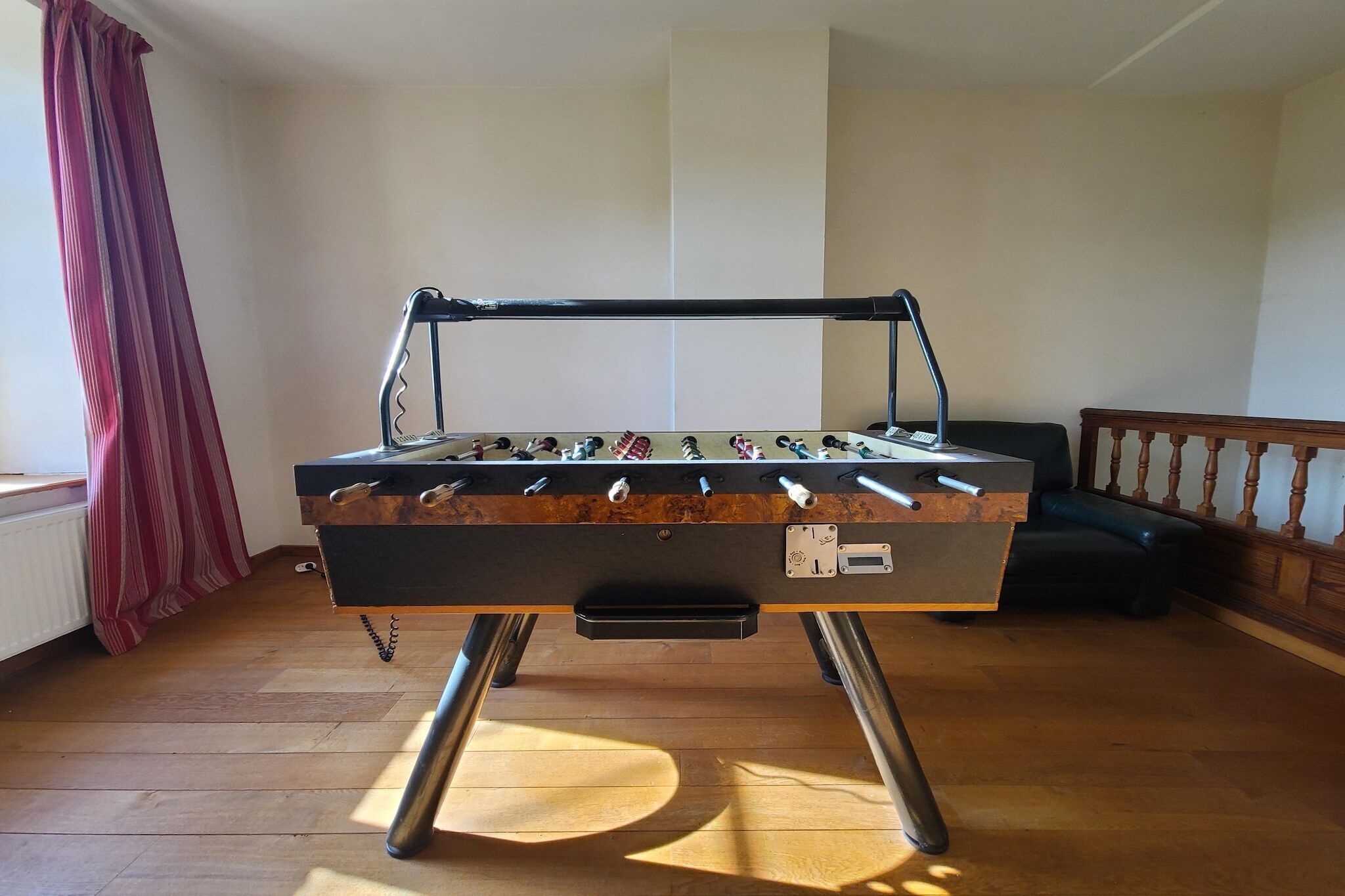 A foosball table in a room with wooden flooring, a window with red curtains, and a black leather sofa.