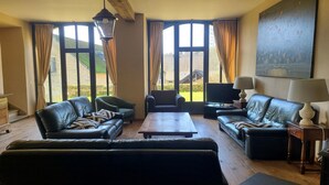 A living room with a sofa, armchairs, a coffee table, and a TV. Large windows with curtains are visible. - Accommodation in the forges of Orval (Orval)