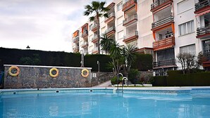 A swimming pool with stone walls and lifebuoys, surrounded by a multi-story building with balconies and palm trees.