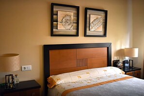 Ein Hotelzimmer mit einem Bett, zwei gerahmten Kunstwerken darüber, einem Nachttisch mit Lampe sowie einem Schreibtisch mit Lampe.