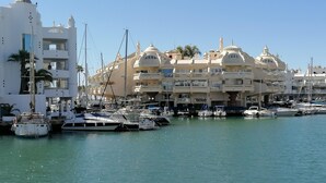 A marina with multiple boats docked, a large white building with balconies, and a clear blue sky.