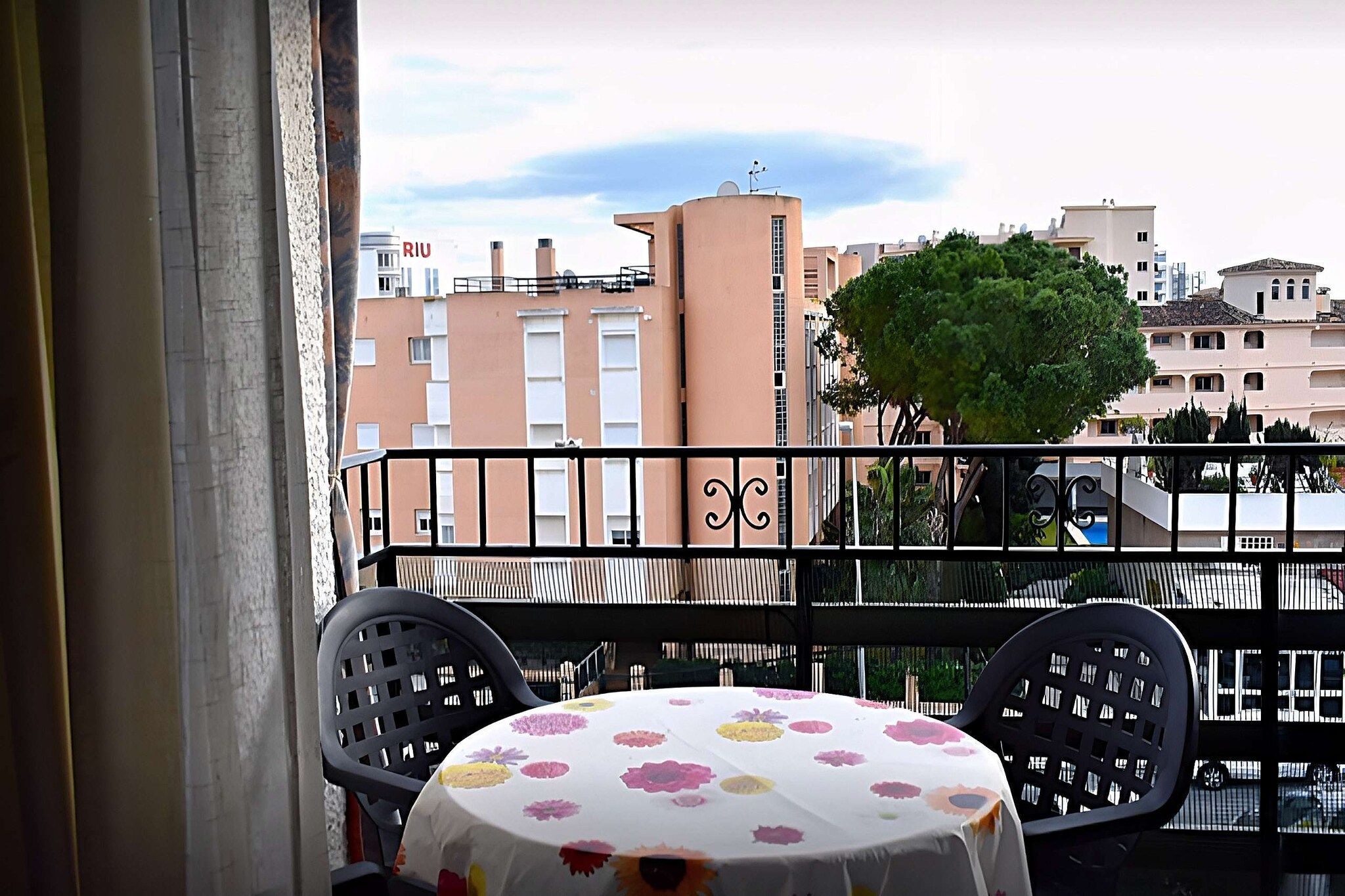 A balcony with a table and chairs, overlooking a residential area with buildings and a tree.