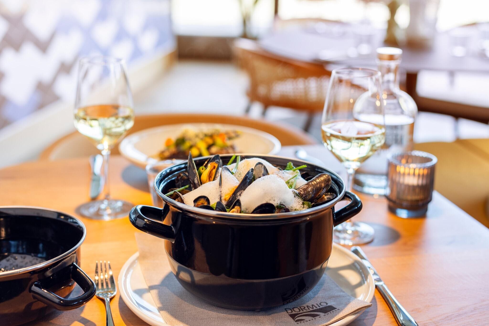 A dining table with a black pot of seafood, a glass of white wine, and a candle.