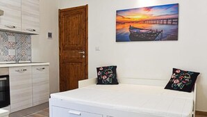 A bedroom with a bed, a wooden door, a kitchen area with patterned tiles, and a sunset painting on the wall.