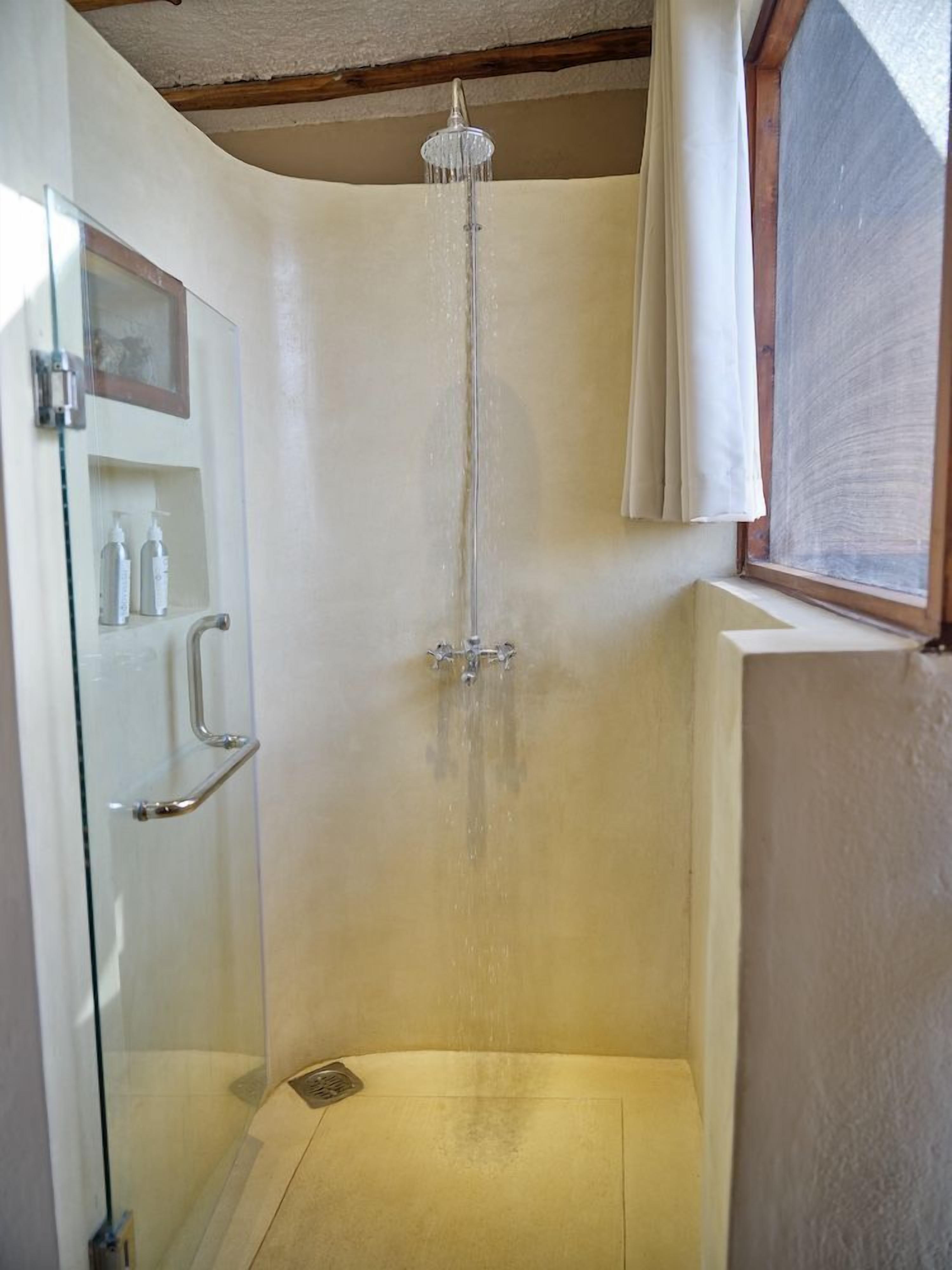 A shower with a glass door, a window with a curtain, and a shelf with toiletries.