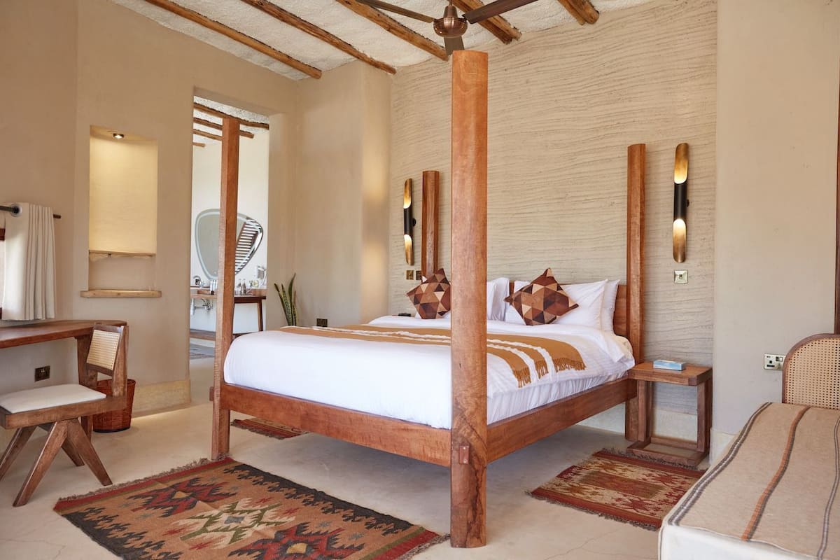 Una cama con dosel y estructura de madera, una mesita de noche con lámpara, una silla y un escritorio con espejo.