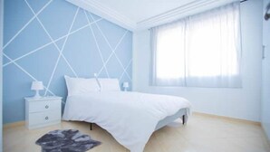 Ett sovrum med en säng, ett sängbord, en lampa, ett fönster med gardiner och en vägg med ett geometriskt mönster.