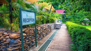 Un área de recepción con una pared de piedra, un cartel que indica el camino a la recepción y un sendero bordeado de follaje.
