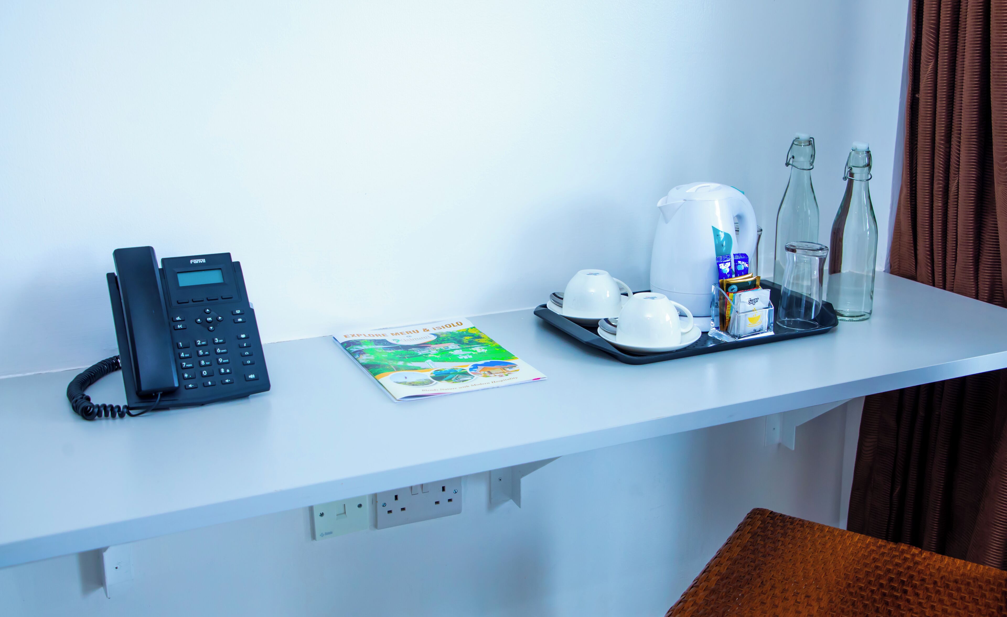 A hotel room desk with a landline phone, a travel brochure, a tray with a coffee maker, cups, a bottle of water, and a glass bottle.