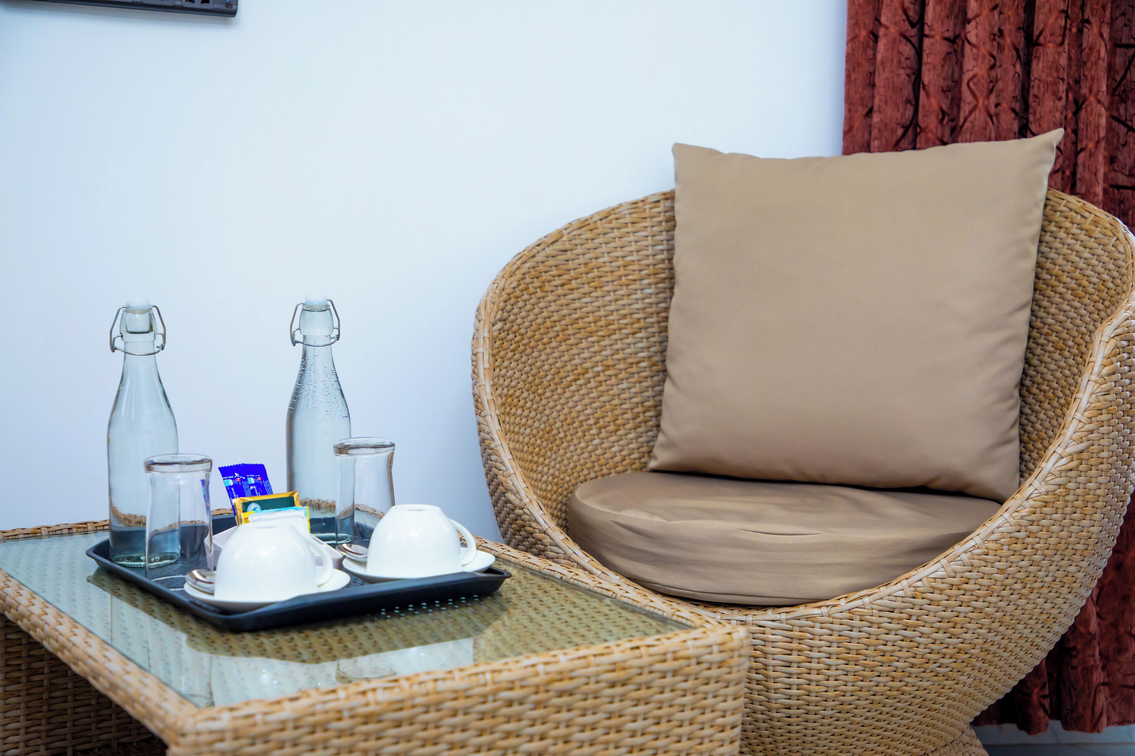 A wicker chair with a beige cushion, a tray with two glass bottles, a white cup, and a blue packet on a glass table.