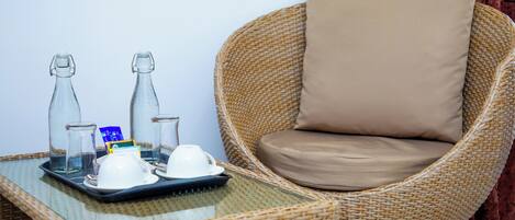 A wicker chair with a beige cushion, a tray with two glass bottles, a white cup, and a blue packet on a glass table.