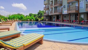 Una piscina con sillones de playa, un edificio de varios pisos con balcones y zonas de estar al aire libre.