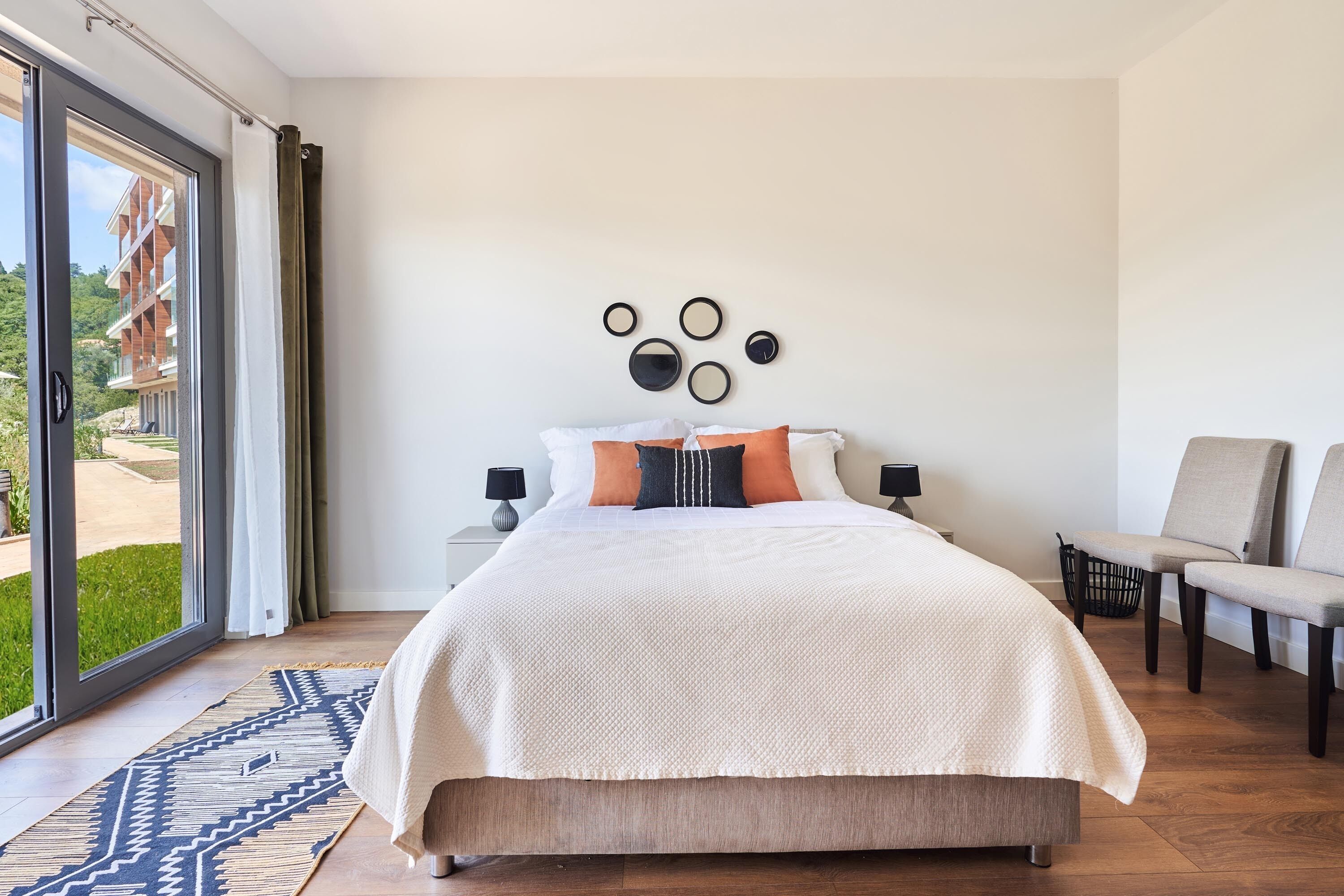 A bedroom with a large bed, two bedside tables, a chair, a patterned rug, and a view through a glass door.