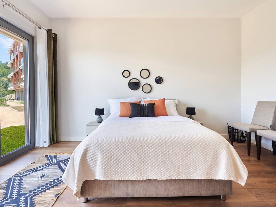 Un dormitorio con una cama grande, dos mesitas de noche, una silla, una alfombra estampada y vistas a través de un ventanal.