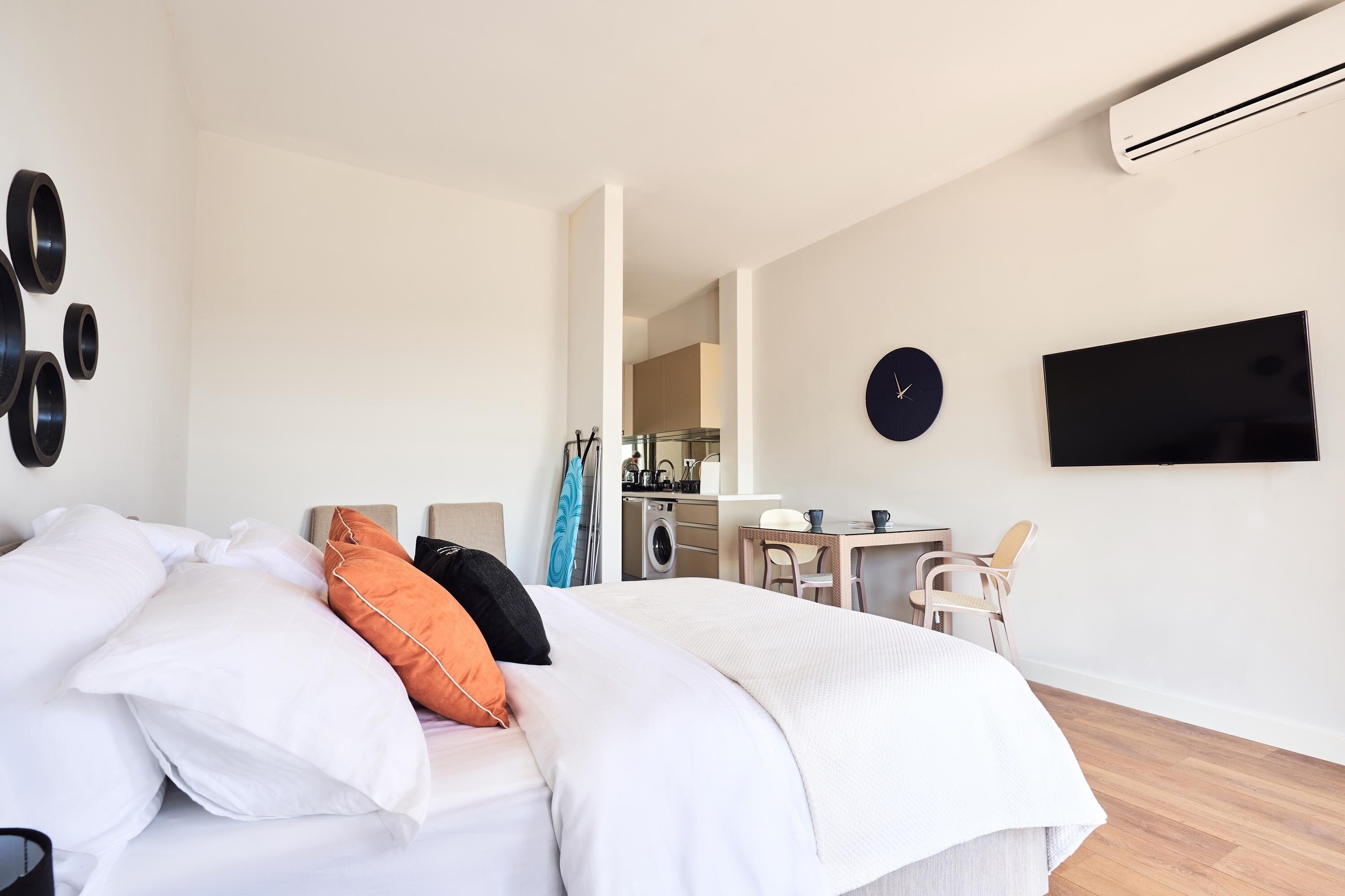 A modern hotel room with a bed, a TV, a dining area, and a kitchen in the background.