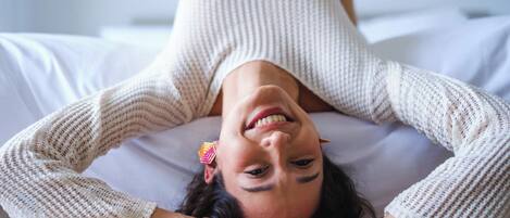 Eine Person liegt mit weißer Bettwäsche auf einem Bett und trägt einen weißen Pullover. Auf dem Arm ist eine Tätowierung sichtbar.