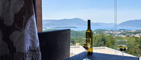 A balcony with a table, a bottle of wine, and two glasses, overlooking a scenic landscape with mountains and a body of water.