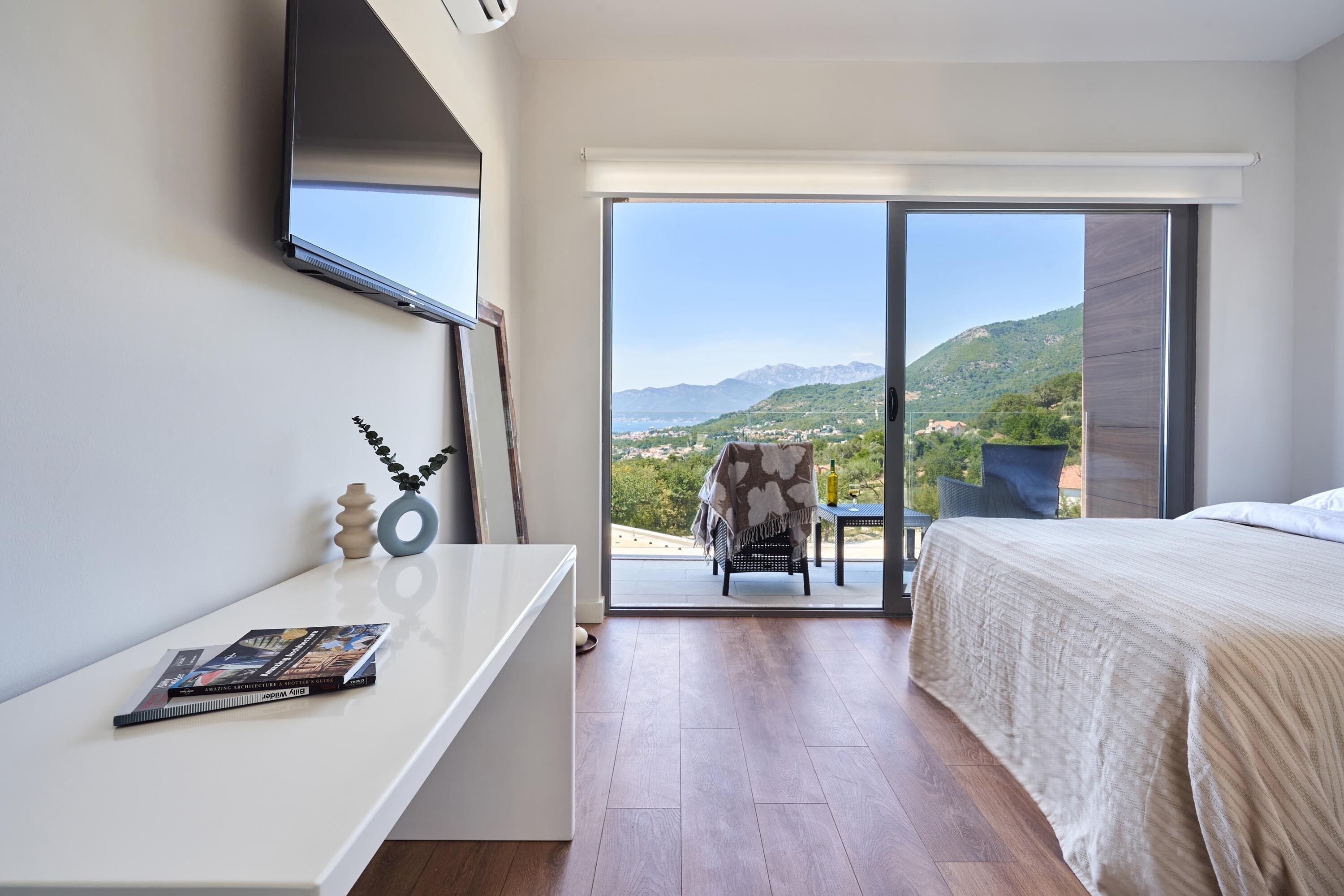 Ein Schlafzimmer mit Bett, Schreibtisch, Stuhl und Blick auf eine gebirgige Landschaft durch eine Schiebetür.