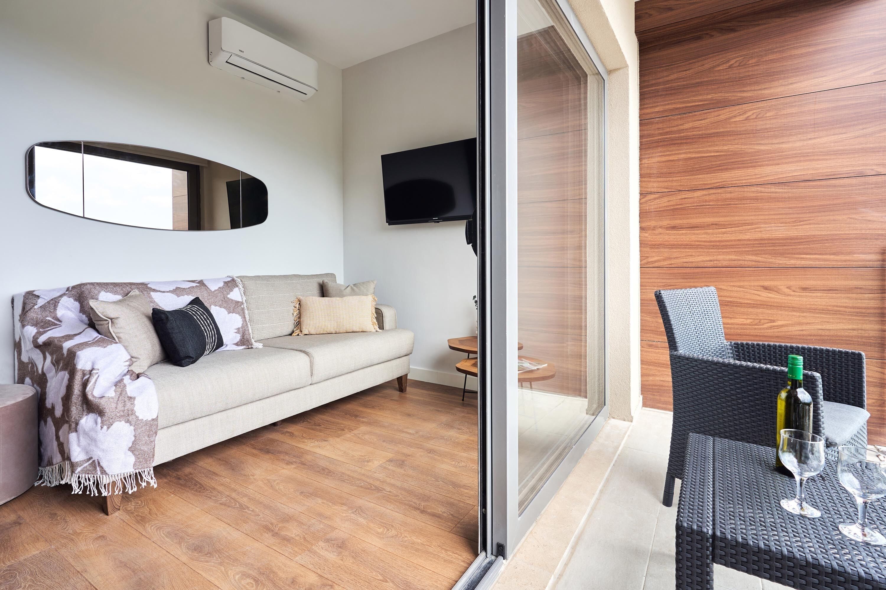 A modern living room with a sofa, a flat-screen TV, and a glass door leading to an outdoor area with a wicker table and chairs.
